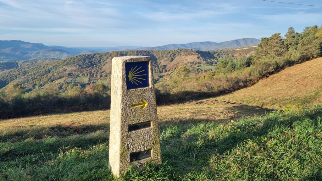Paaltje met symbool van camino en gele pijl op de voorgrond, prachtig uitzicht erachter.